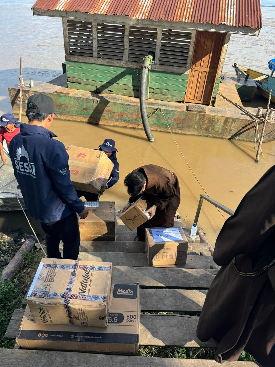 Entrega de caixas e suprimentos em passarela de madeira à margem do rio.