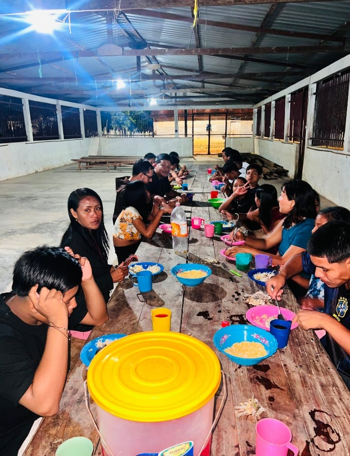 Jovens e frades partilhando a refeição em longa mesa de madeira.