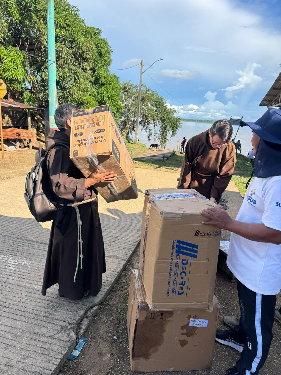 Frades e agentes descarregando caixas de doações na beira do rio.