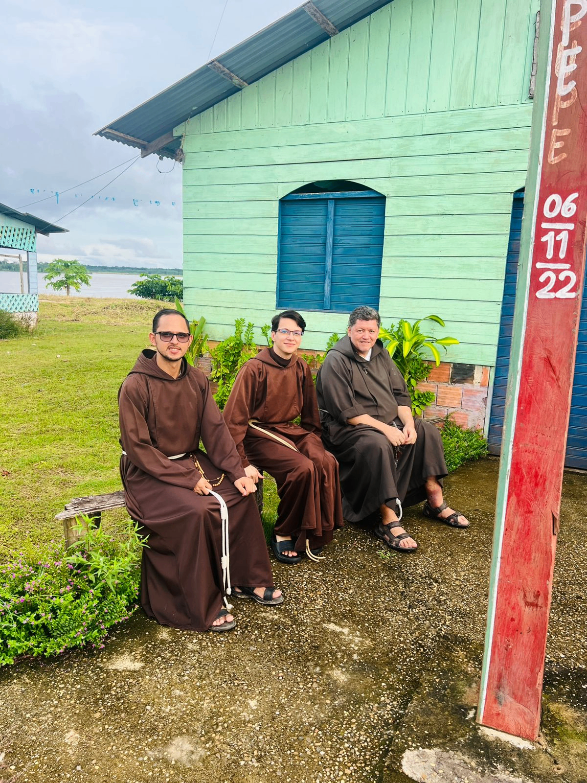 Três frades capuchinhos sentados em frente a casa ribeirinha à beira do rio